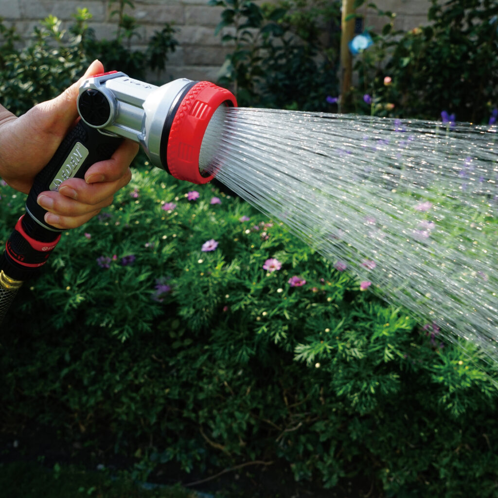 Hand holding the Eden Metal Shower Nozzle and watering in front of green bushes.